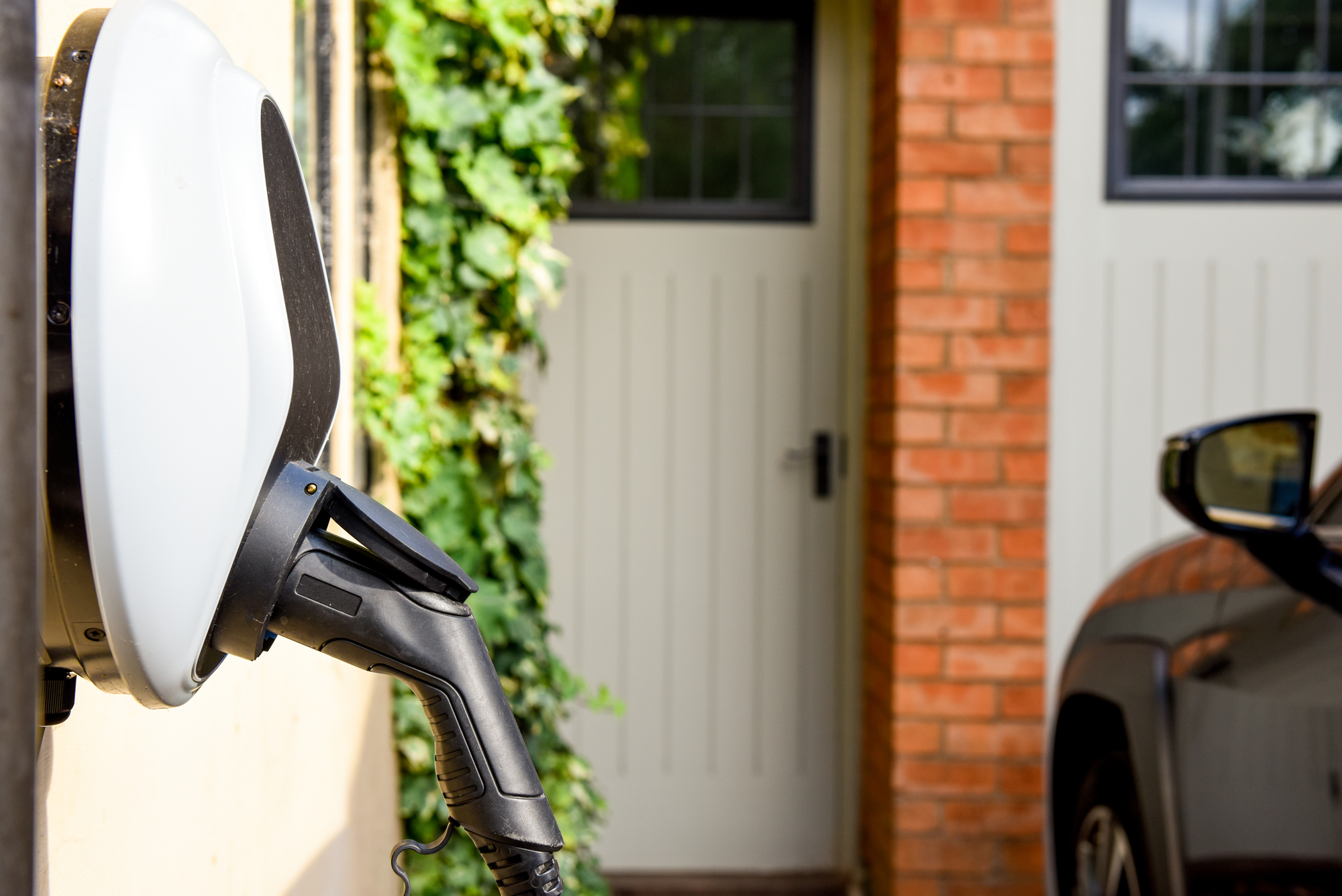 An electric vehicle charger is plugged into the wall outside a home, with a partial view of a car on the right and a brick house with a garage door and greenery in the background